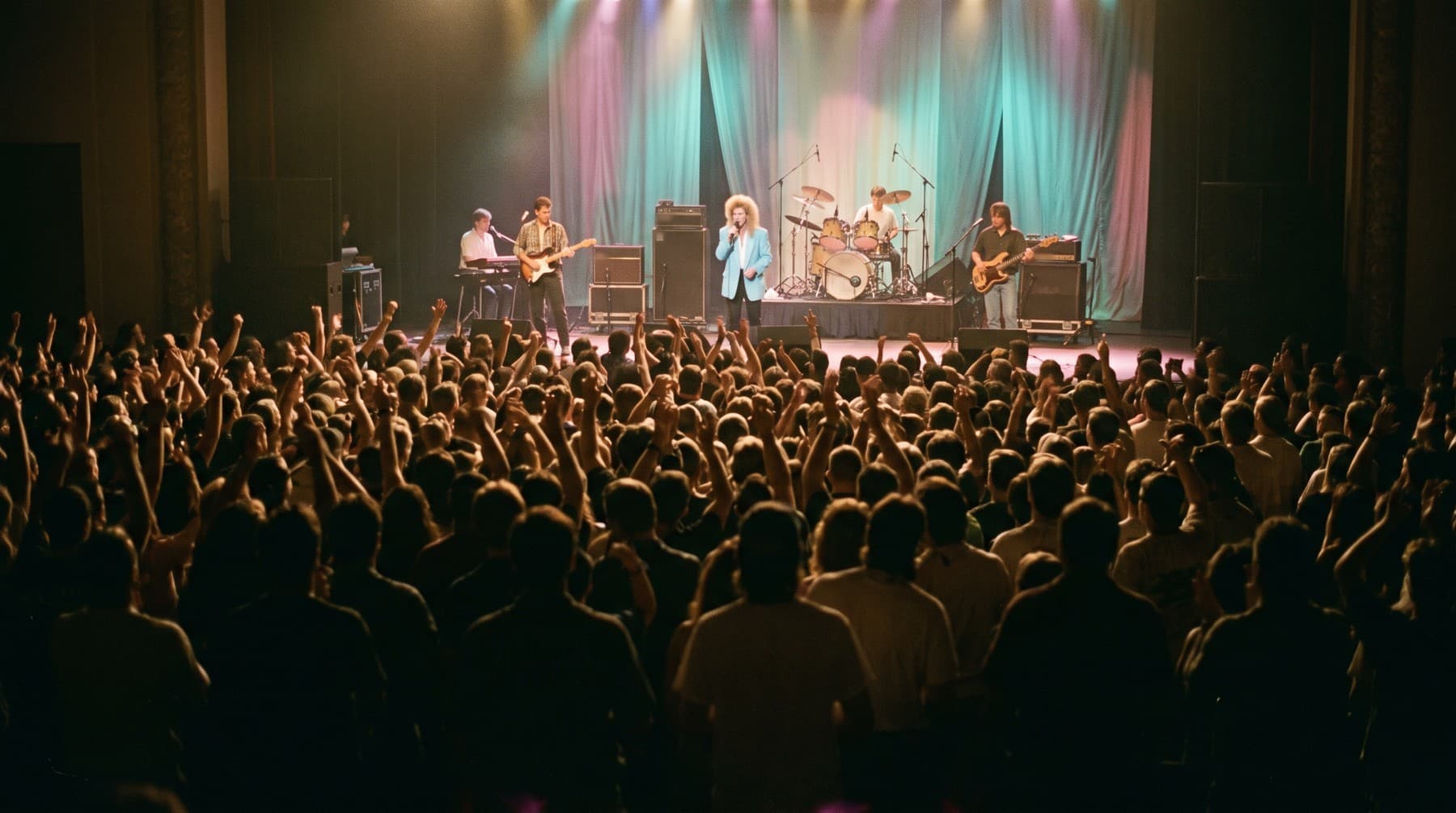A packed hall seen from the back, hands raised toward a lit stage where a five-piece band plays in front of teal and pink curtains